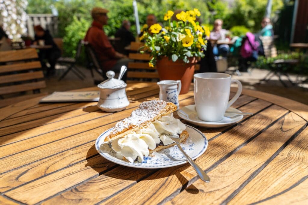 Angeschnittene Friesentorte mit Sahne und Puderzucker auf einem Holztisch im Gartencafé in Wyk, daneben eine Tasse Kaffee.