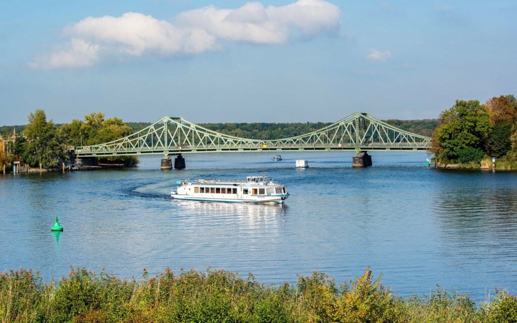 Die Glienicker Brücke über die Havel zwischen Potsdam und Berlin, davor ein weisses Ausflugsschiff der Weissen Flotte auf dem Wasser unter blauem Himmel.