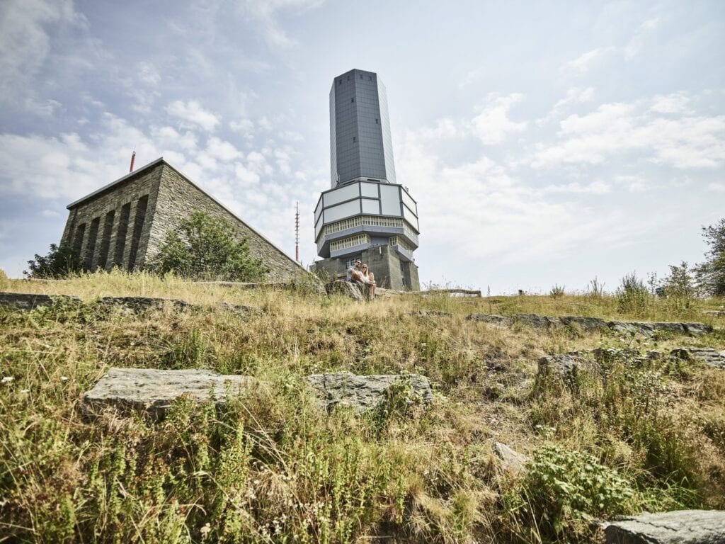 Der Fernmeldeturm auf dem Großen Feldberg im Taunus, davor ein Paar, das auf Felsen in einer sommerlichen Wiese sitzt, im Hintergrund das steinerne Feldberghaus.