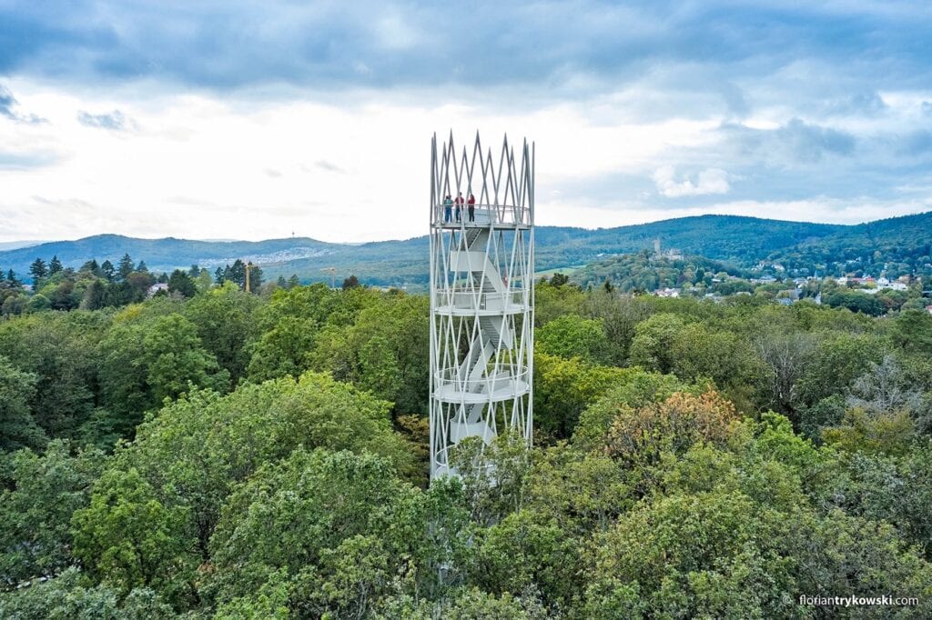 Der weiße stählerne Hardtbergturm mit gewendelter Treppe ragt aus dem Wald im Taunus, oben stehen drei Personen auf der Aussichtsplattform, in der Ferne die Burg Kronberg.
