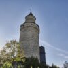 Der runde steinerne Hexenturm in Idstein im Taunus von unten gesehen, daneben tief stehende Sonne und ein angrenzendes Fachwerkgebäude.