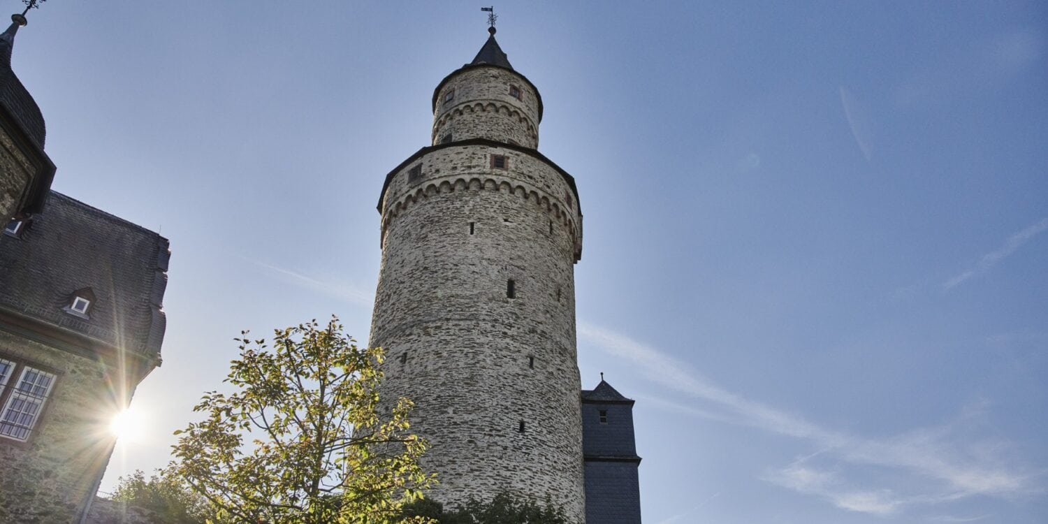 Der runde steinerne Hexenturm in Idstein im Taunus von unten gesehen, daneben tief stehende Sonne und ein angrenzendes Fachwerkgebäude.