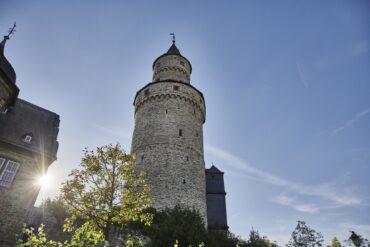 Der runde steinerne Hexenturm in Idstein im Taunus von unten gesehen, daneben tief stehende Sonne und ein angrenzendes Fachwerkgebäude.