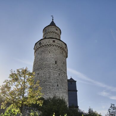 Der runde steinerne Hexenturm in Idstein im Taunus von unten gesehen, daneben tief stehende Sonne und ein angrenzendes Fachwerkgebäude.