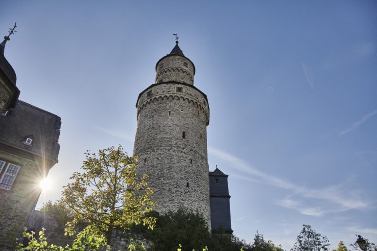 Der runde steinerne Hexenturm in Idstein im Taunus von unten gesehen, daneben tief stehende Sonne und ein angrenzendes Fachwerkgebäude.