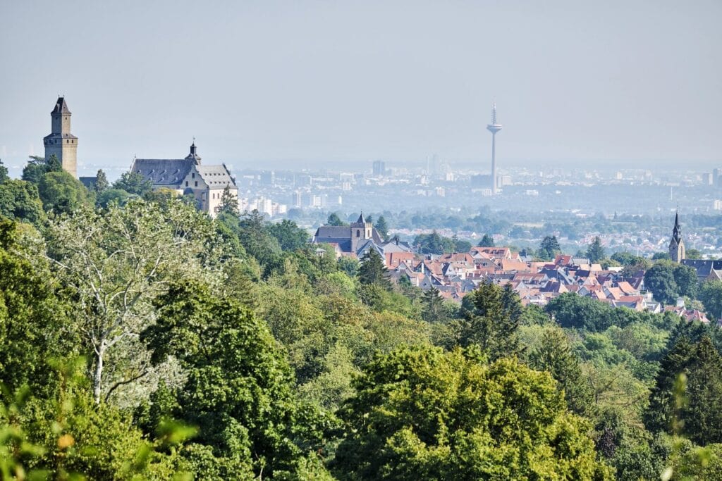 Blick vom Malerblick in Kronberg im Taunus über die Baumwipfel auf die Burg Kronberg links und die im Dunst liegende Skyline von Frankfurt mit dem Europaturm.