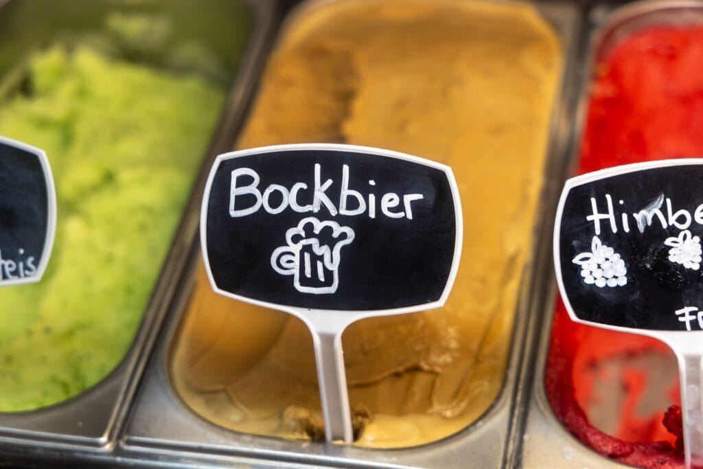 Eistheke mit einer Sorte Bockbiereis, davor steckt ein Schild mit der Aufschrift Bockbier und einer gemalten Bierkrug-Skizze. Links und rechts stehen Behälter mit grünem und rotem Eis.