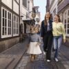 Eine als Katze verkleidete Figur in Pullover und Faltenrock, die zusammen mit zwei lachenden Frauen eine Kopfsteinpflastergasse im Schnoor-Viertel entlanggeht.