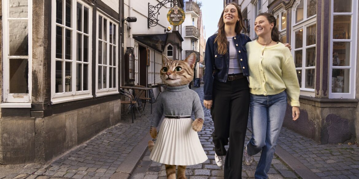 Eine als Katze verkleidete Figur in Pullover und Faltenrock, die zusammen mit zwei lachenden Frauen eine Kopfsteinpflastergasse im Schnoor-Viertel entlanggeht.