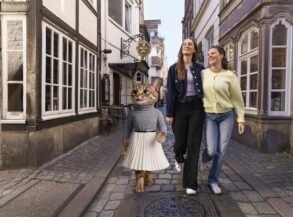 Eine als Katze verkleidete Figur in Pullover und Faltenrock, die zusammen mit zwei lachenden Frauen eine Kopfsteinpflastergasse im Schnoor-Viertel entlanggeht.