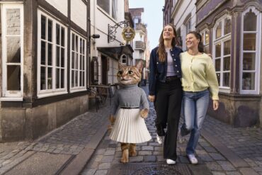 Eine als Katze verkleidete Figur in Pullover und Faltenrock, die zusammen mit zwei lachenden Frauen eine Kopfsteinpflastergasse im Schnoor-Viertel entlanggeht.