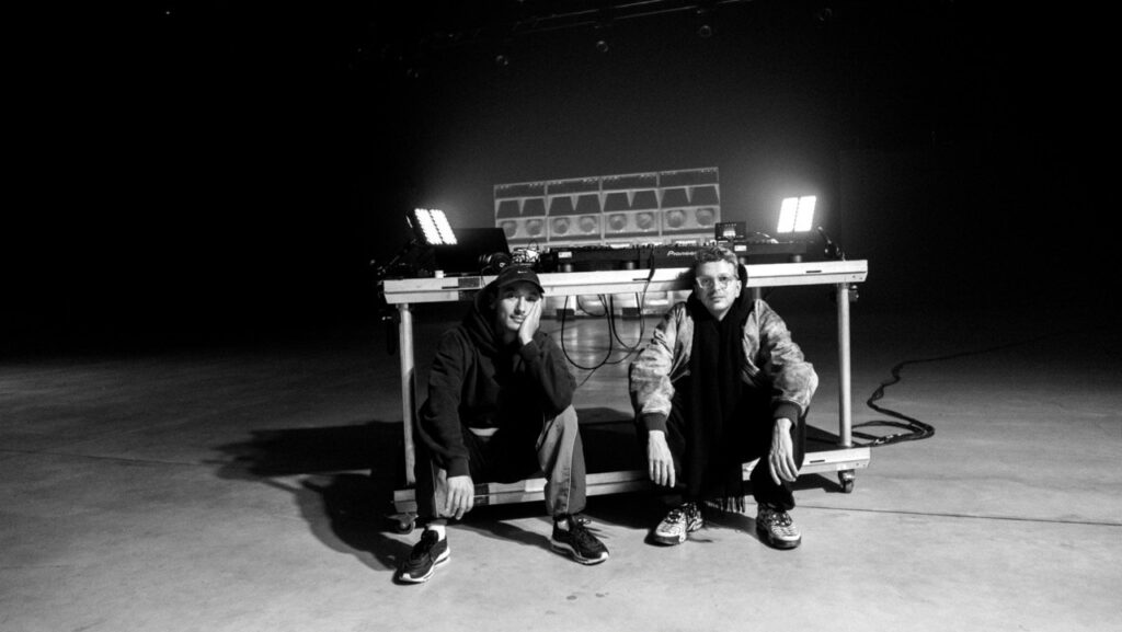 Schwarzweißes Pressefoto zum Album „Kitschkrieg Zwei": zwei Musiker hocken vor einem rollbaren DJ-Pult mit Plattenspielern und Lautsprecherboxen in einer leeren Halle.