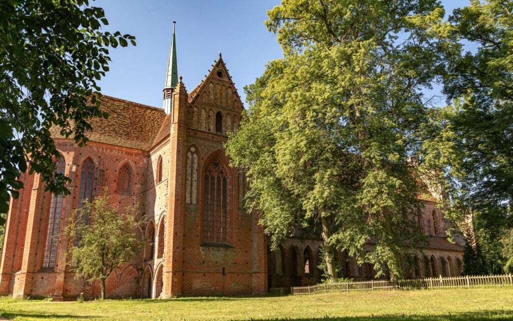Das gotische Backsteinkloster Chorin in Brandenburg mit verzierter Giebelfassade und schlankem Dachreiter, umgeben von Bäumen und Wiese unter blauem Himmel.