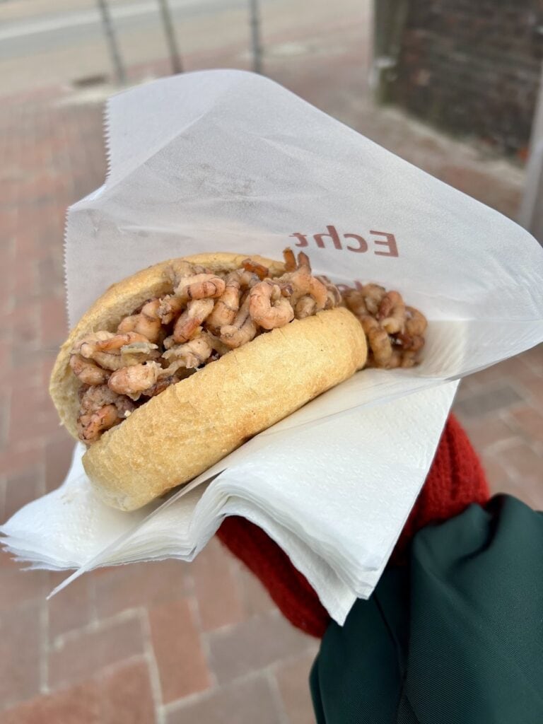Hand hält ein Krabbenbrötchen mit Nordseekrabben in einer Papiertüte über gepflastertem Boden.