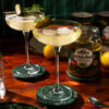 Two glasses of martini, garnished with a pickle. In the background stands an opened glass of pickles.