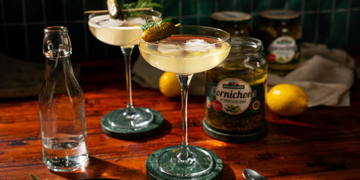 Two glasses of martini, garnished with a pickle. In the background stands an opened glass of pickles.