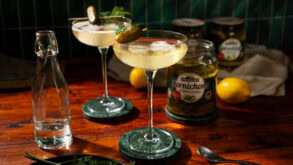Two glasses of martini, garnished with a pickle. In the background stands an opened glass of pickles.