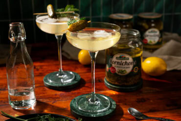 Two glasses of martini, garnished with a pickle. In the background stands an opened glass of pickles.