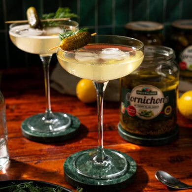 Two glasses of martini, garnished with a pickle. In the background stands an opened glass of pickles.
