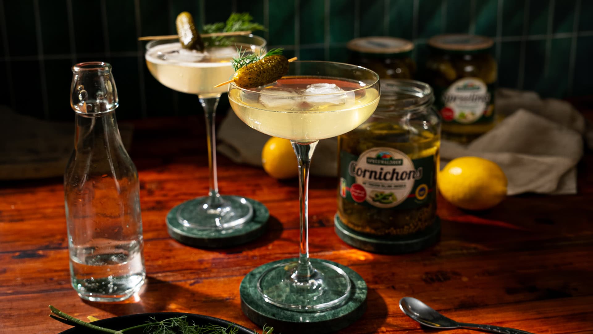 Two glasses of martini, garnished with a pickle. In the background stands an opened glass of pickles.
