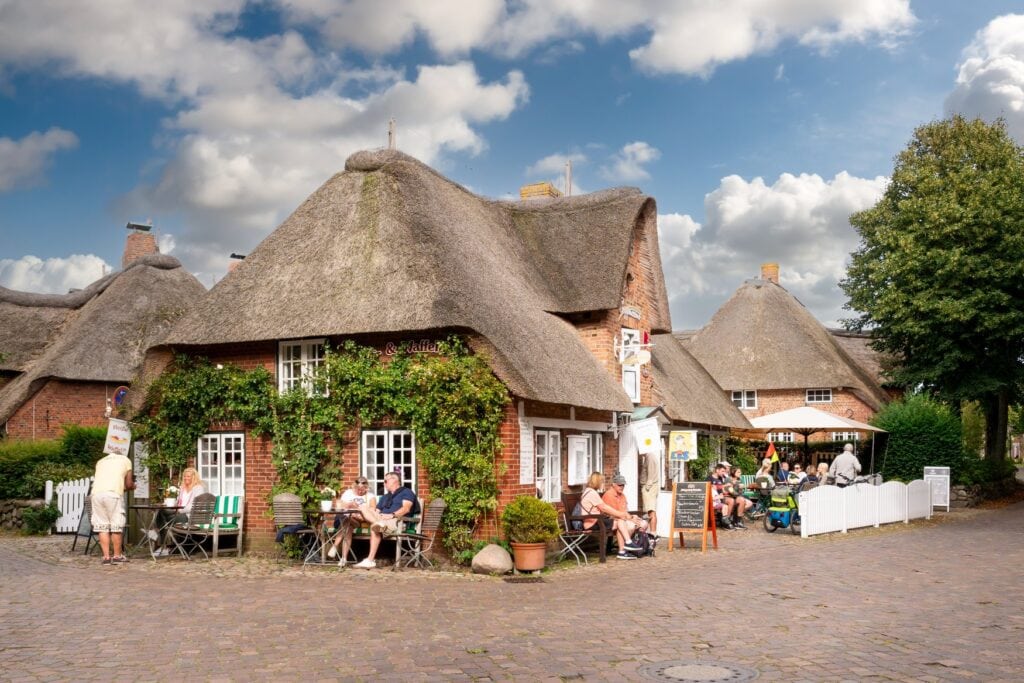 Reetgedecktes Backsteinhaus mit Café und Außenplätzen im Ortskern von Nieblum, davor Kopfsteinpflaster und Gäste an den Tischen.