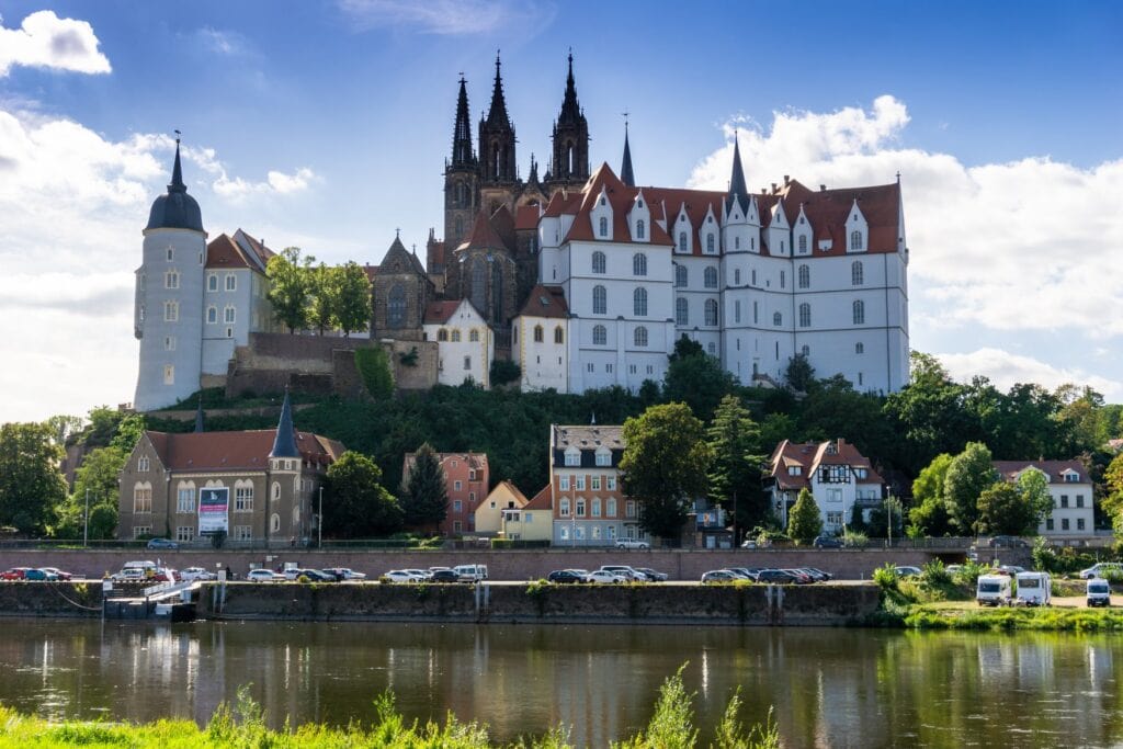 Blick über die Elbe auf den Burgberg von Meissen mit der Albrechtsburg und den Türmen des Doms, darunter Wohnhäuser und eine Uferpromenade mit Parkplätzen.