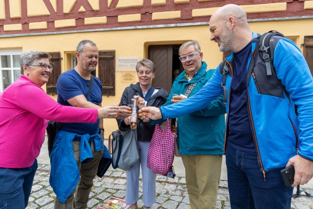 Fünf Teilnehmende einer Stadtführung in Hersbruck stoßen mit kleinen Gläsern eines Hopfenlikörs an. Sie stehen vor einem gelben Fachwerkhaus, auf dem Boden liegt eine Flasche mit Etikett.