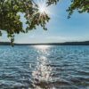 Blick durch überhängende Blätter auf den Stechlinsee in Brandenburg, die Sonne steht als Stern zwischen den Zweigen und glitzert auf dem Wasser, am Horizont ein bewaldetes Ufer.