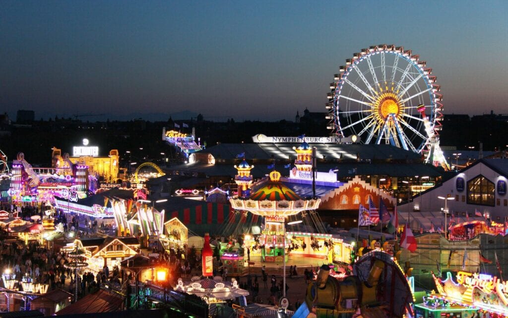 Blick über die beleuchtete Theresienwiese bei Nacht mit Riesenrad, Karussells und Festzelten.