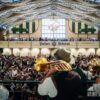 Blasmusiker mit Trompeten und Tuba spielen von der Bühne in ein volles Festzelt mit Galerie und Zeltdekoration.
