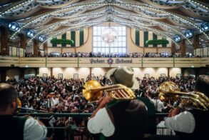 Blasmusiker mit Trompeten und Tuba spielen von der Bühne in ein volles Festzelt mit Galerie und Zeltdekoration.