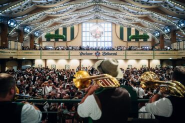 Blasmusiker mit Trompeten und Tuba spielen von der Bühne in ein volles Festzelt mit Galerie und Zeltdekoration.