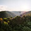 Ein hölzerner Aussichtspavillon steht auf einer bewaldeten Kuppe über dem Rheintal, darüber ziehen die Sessel der Bopparder Sesselbahn ihre Bahn. Die tiefstehende Sonne liegt im Dunst über den Höhenzügen.
