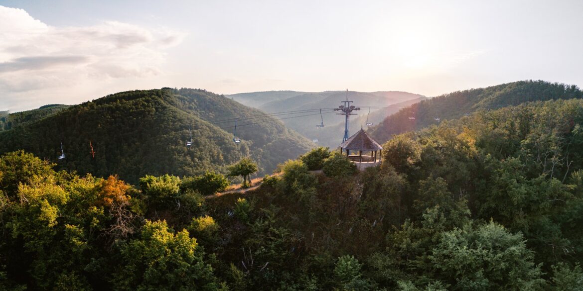 Ein hölzerner Aussichtspavillon steht auf einer bewaldeten Kuppe über dem Rheintal, darüber ziehen die Sessel der Bopparder Sesselbahn ihre Bahn. Die tiefstehende Sonne liegt im Dunst über den Höhenzügen.