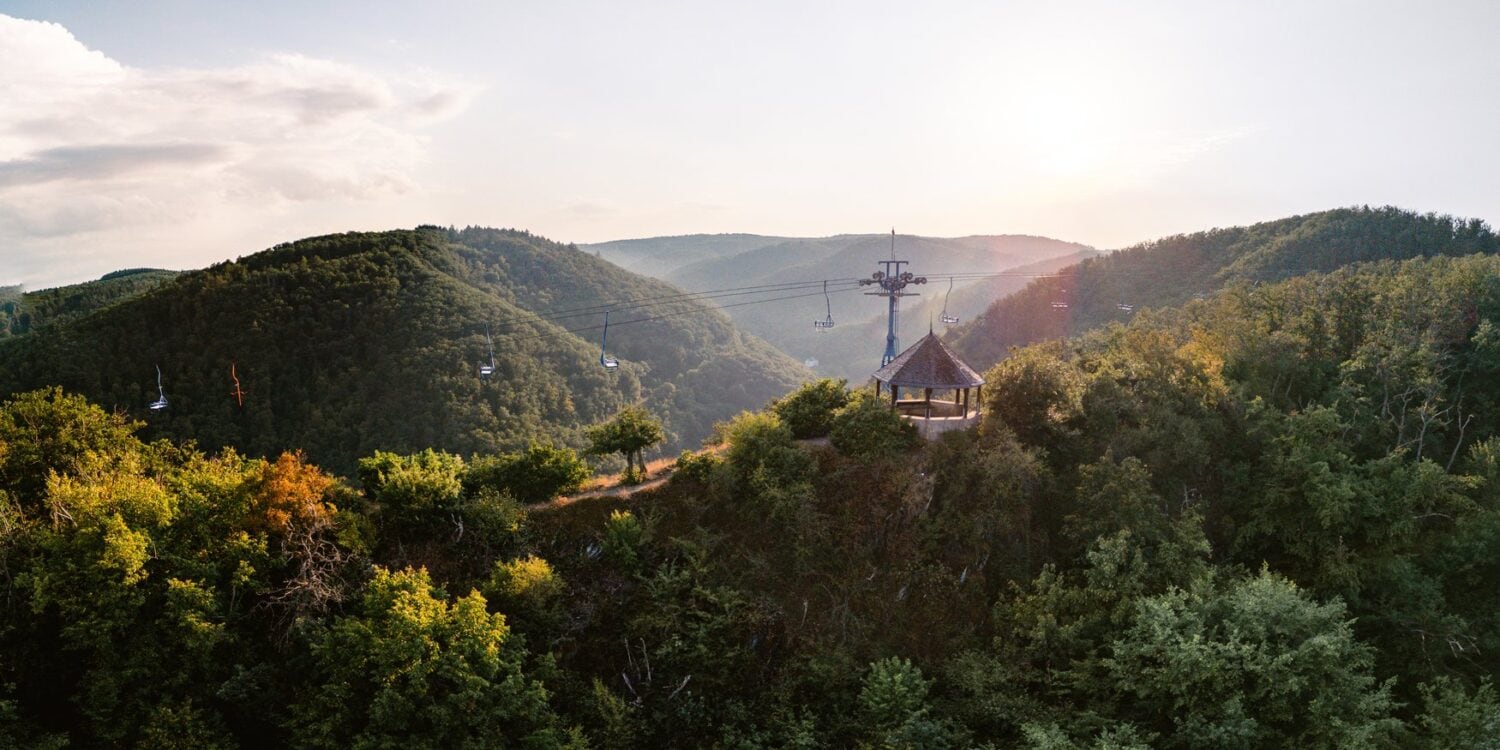 Ein hölzerner Aussichtspavillon steht auf einer bewaldeten Kuppe über dem Rheintal, darüber ziehen die Sessel der Bopparder Sesselbahn ihre Bahn. Die tiefstehende Sonne liegt im Dunst über den Höhenzügen.