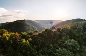 Ein hölzerner Aussichtspavillon steht auf einer bewaldeten Kuppe über dem Rheintal, darüber ziehen die Sessel der Bopparder Sesselbahn ihre Bahn. Die tiefstehende Sonne liegt im Dunst über den Höhenzügen.