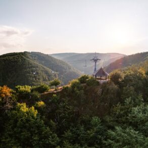 Ein hölzerner Aussichtspavillon steht auf einer bewaldeten Kuppe über dem Rheintal, darüber ziehen die Sessel der Bopparder Sesselbahn ihre Bahn. Die tiefstehende Sonne liegt im Dunst über den Höhenzügen.