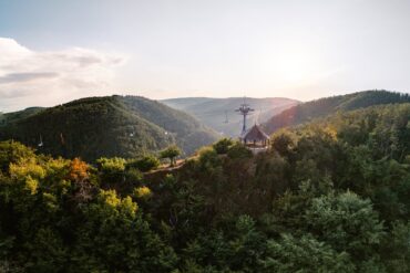 Ein hölzerner Aussichtspavillon steht auf einer bewaldeten Kuppe über dem Rheintal, darüber ziehen die Sessel der Bopparder Sesselbahn ihre Bahn. Die tiefstehende Sonne liegt im Dunst über den Höhenzügen.
