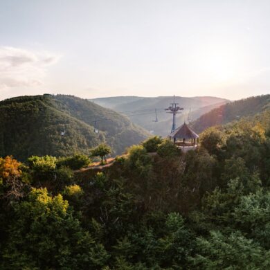 Ein hölzerner Aussichtspavillon steht auf einer bewaldeten Kuppe über dem Rheintal, darüber ziehen die Sessel der Bopparder Sesselbahn ihre Bahn. Die tiefstehende Sonne liegt im Dunst über den Höhenzügen.
