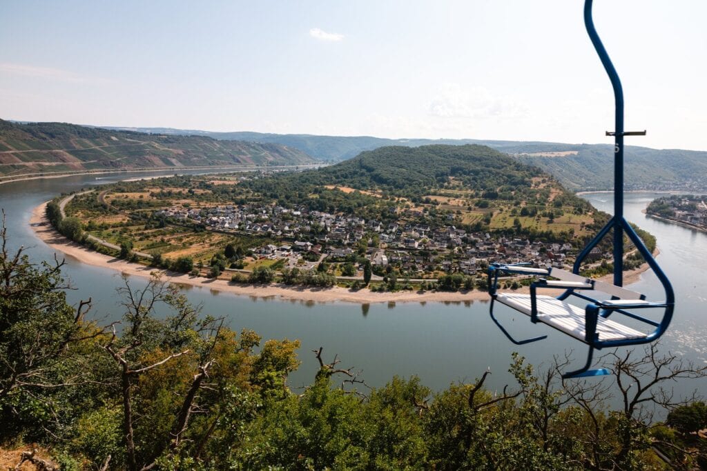 Ein leerer blauer Sessel der Bopparder Sesselbahn schwebt über dem Hang, darunter liegt die Rheinschleife mit dem Ort Filsen auf der Halbinsel und Weinbergen am gegenüberliegenden Ufer.