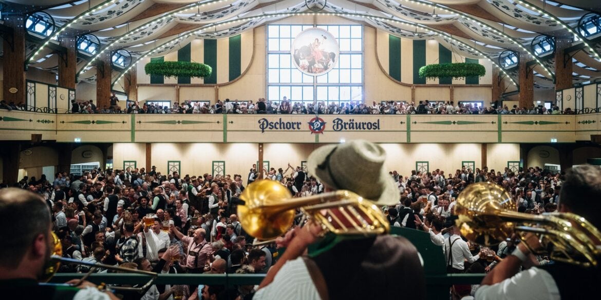 Brass musicians with trumpets and a tuba play from the stage into a packed beer tent with a gallery above.