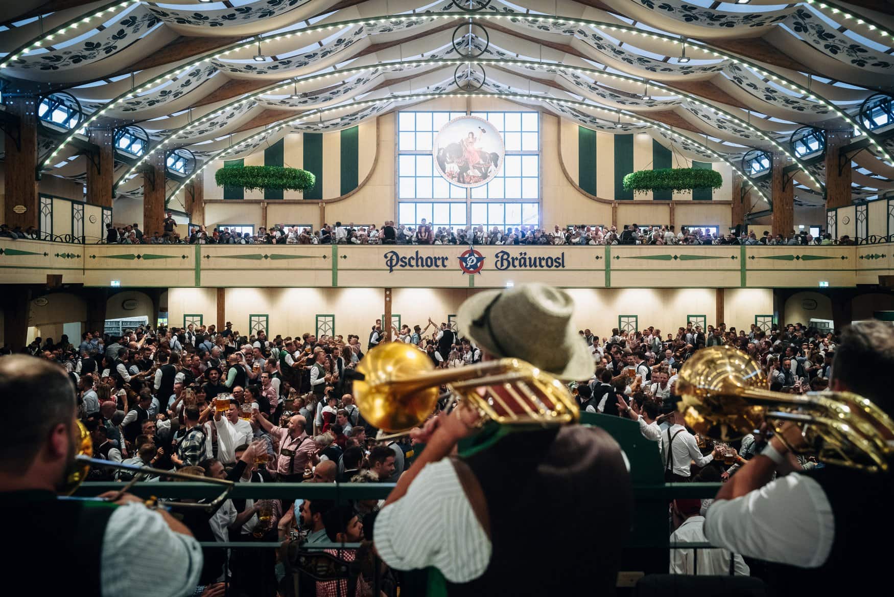 Brass musicians with trumpets and a tuba play from the stage into a packed beer tent with a gallery above.