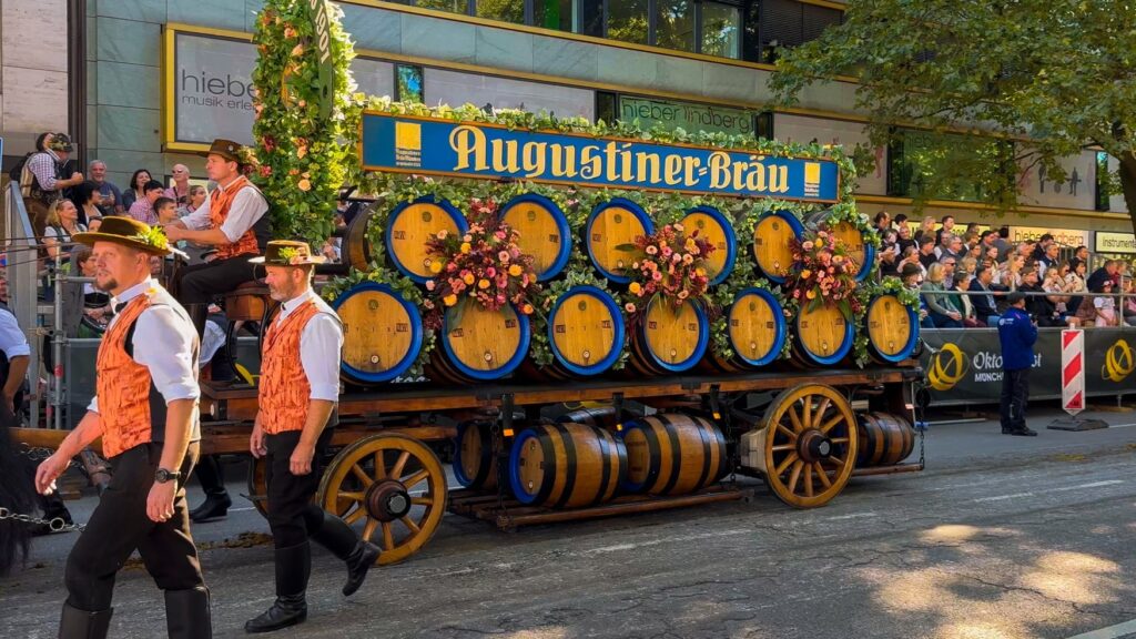 Mit Blumen geschmückter Bierwagen von Augustiner-Bräu beim Wiesneinzug, daneben Männer in orangefarbenen Westen.