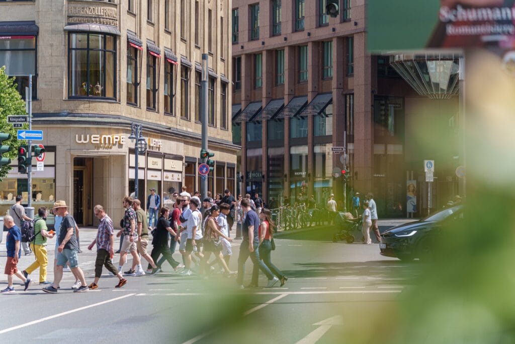 Fußgänger überqueren eine Straße an der Königsallee in Düsseldorf, links das Hohenzollernhaus mit Wempe-Geschäft, rechts Ladenlokale von Sephora und Repeat.