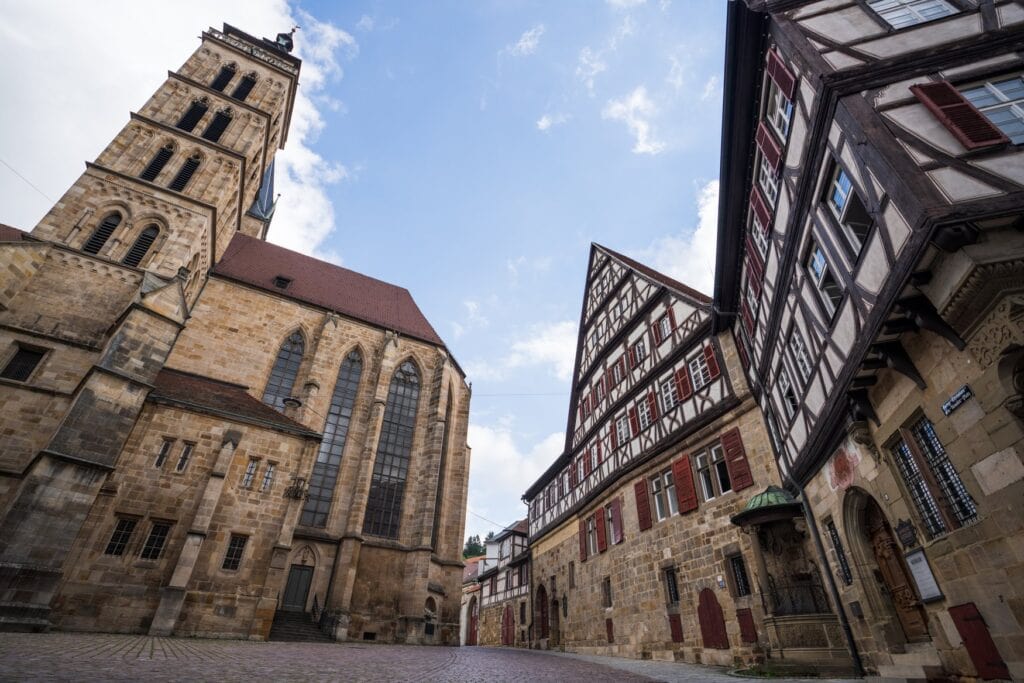 Untersicht auf die Stadtkirche St. Dionys in der Esslinger Altstadt, rechts daneben ein Fachwerkhaus mit roten Fensterläden über einem Sockel aus Sandstein. Der Platz davor ist mit Kopfstein gepflastert.