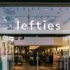 Glasfassade einer Lefties-Filiale in Badalona bei Barcelona mit beleuchtetem Schriftzug über dem Eingang, im Schaufenster hängen Plakate mit Kindermode, dahinter der Verkaufsraum.