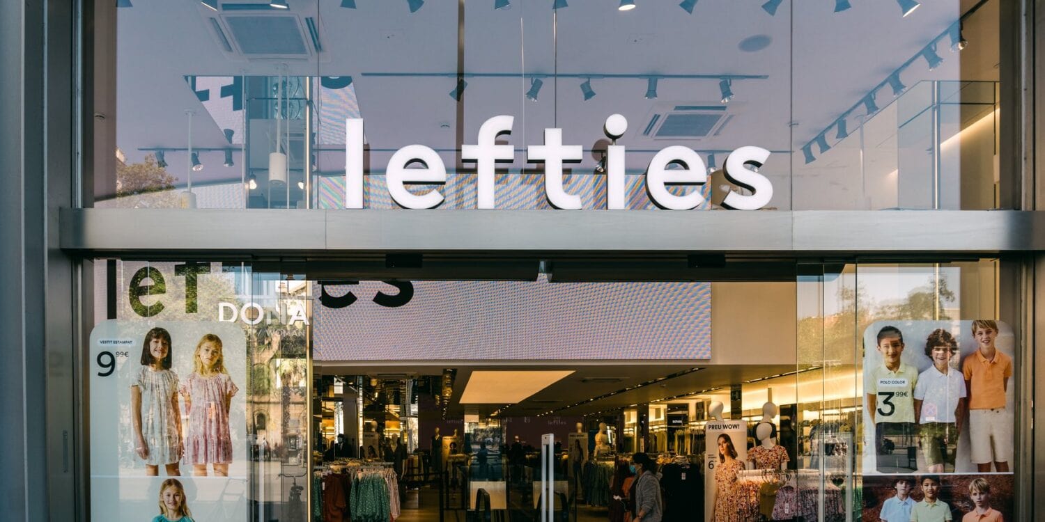 Glasfassade einer Lefties-Filiale in Badalona bei Barcelona mit beleuchtetem Schriftzug über dem Eingang, im Schaufenster hängen Plakate mit Kindermode, dahinter der Verkaufsraum.