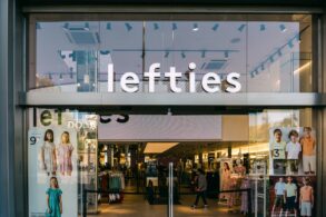 Glasfassade einer Lefties-Filiale in Badalona bei Barcelona mit beleuchtetem Schriftzug über dem Eingang, im Schaufenster hängen Plakate mit Kindermode, dahinter der Verkaufsraum.