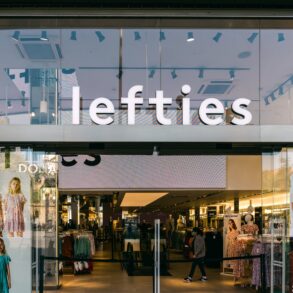 Glasfassade einer Lefties-Filiale in Badalona bei Barcelona mit beleuchtetem Schriftzug über dem Eingang, im Schaufenster hängen Plakate mit Kindermode, dahinter der Verkaufsraum.
