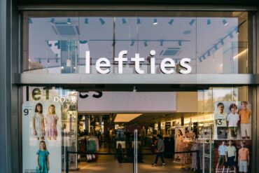 Glasfassade einer Lefties-Filiale in Badalona bei Barcelona mit beleuchtetem Schriftzug über dem Eingang, im Schaufenster hängen Plakate mit Kindermode, dahinter der Verkaufsraum.
